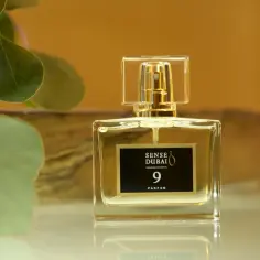 Eleganckie perfumy damskie z Dubaju, Sense Dubai 9 Parfum, w szklanym flakonie z złotymi detalami, otoczone zielonymi liśćmi.