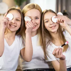 Mama i córki cieszą się pierwszymi kosmetykami dla 12 latki. Uśmiechnięte dziewczynki z wacikami na oczach i mamą z pędzelkiem.