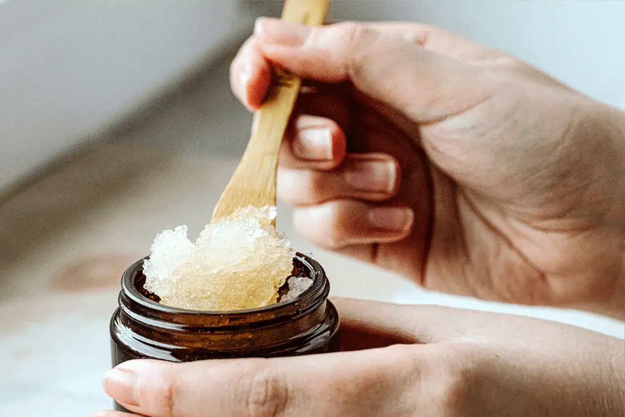 Dłonie nabierają drewnianą łyżeczką złocisty peeling do ciała z małego słoiczka.