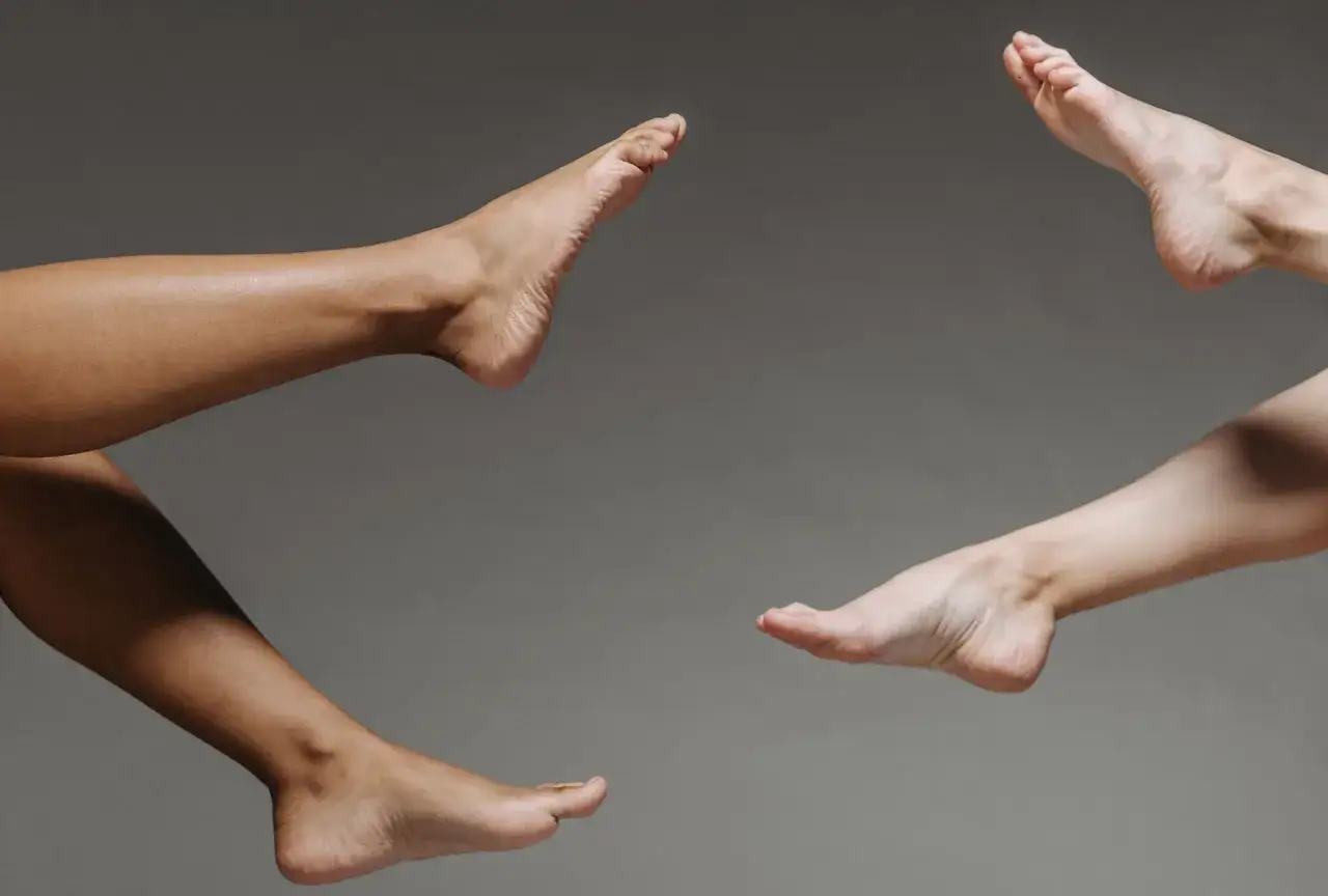 Dwie pary stóp, jedna ciemniejsza, druga jaśniejsza, unoszą się w powietrzu. Klasyczny pedicure co to? To dbałość o piękno stóp.