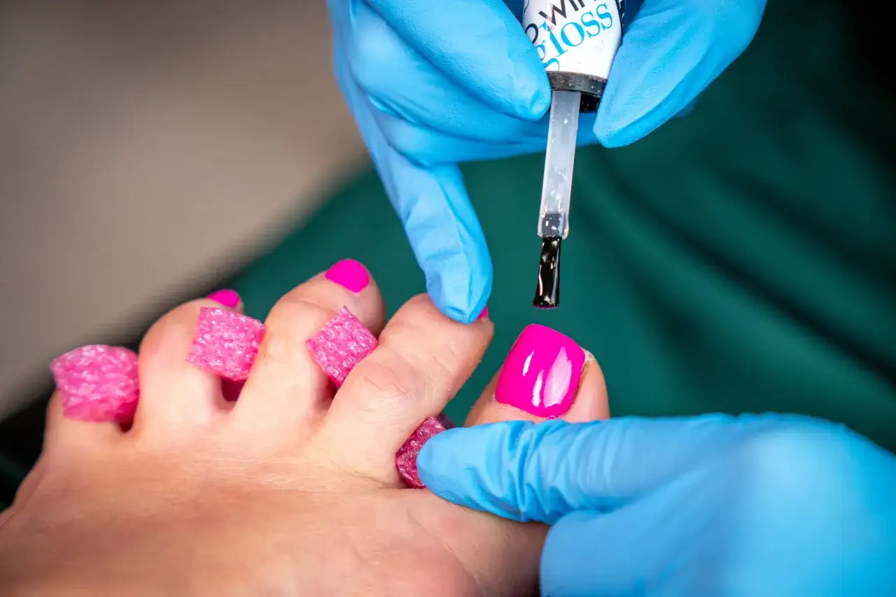Dłonie w niebieskich rękawiczkach aplikują czarny lakier na paznokcie u stóp. To klasyczny pedicure, który odświeża wygląd stóp.
