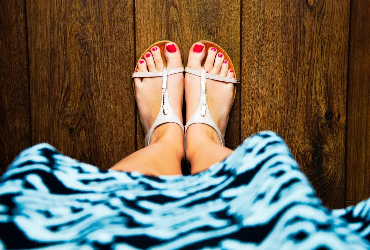 Zadbany pedicure klasyczny co to? Czerwone paznokcie u stóp w sandałkach na drewnianej podłodze.